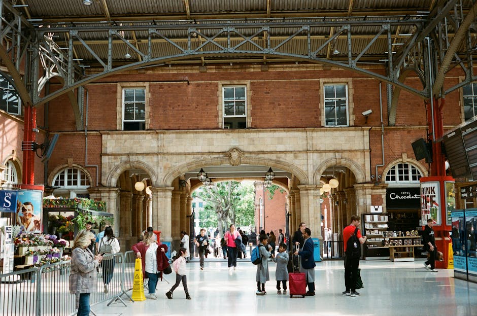The image depicts the interior of a spacious public area along a platform, with a high arched ceiling supported by metal beams and large glass panels allowing natural light to illuminate the space. On