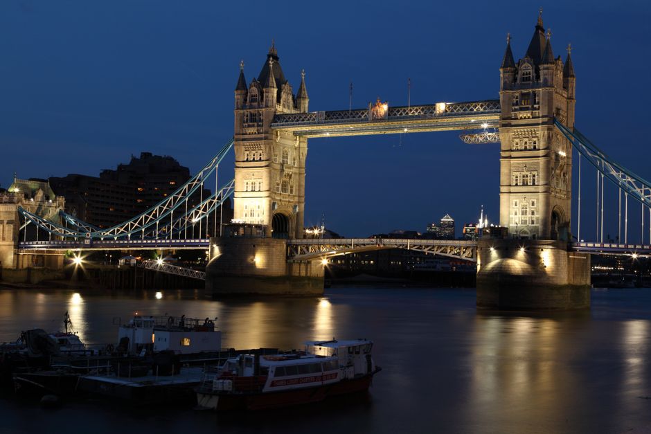 The image displays London's Tower Bridge illuminated at night, with its distinctive twin towers and suspension design reflected in the calm River Thames below. The bridge’s intricate stonework and m