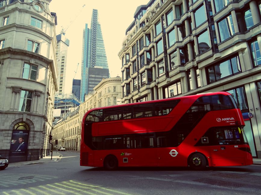 A modern red double-decker bus parked on a city street in central London, surrounded by tall contemporary and historic buildings with glass windows and decorative stone facades. The scene is illuminat